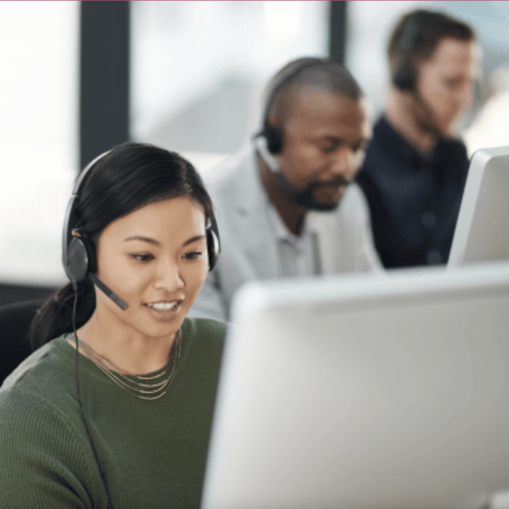 A woman with headphones and a microphone headset is smiling as she looks at a computer screen, likely engaged in a customer service task. She is wearing a green top and a layered necklace. In the background, two male colleagues are also wearing headsets and working on their computers, suggesting a professional office environment, possibly a call center